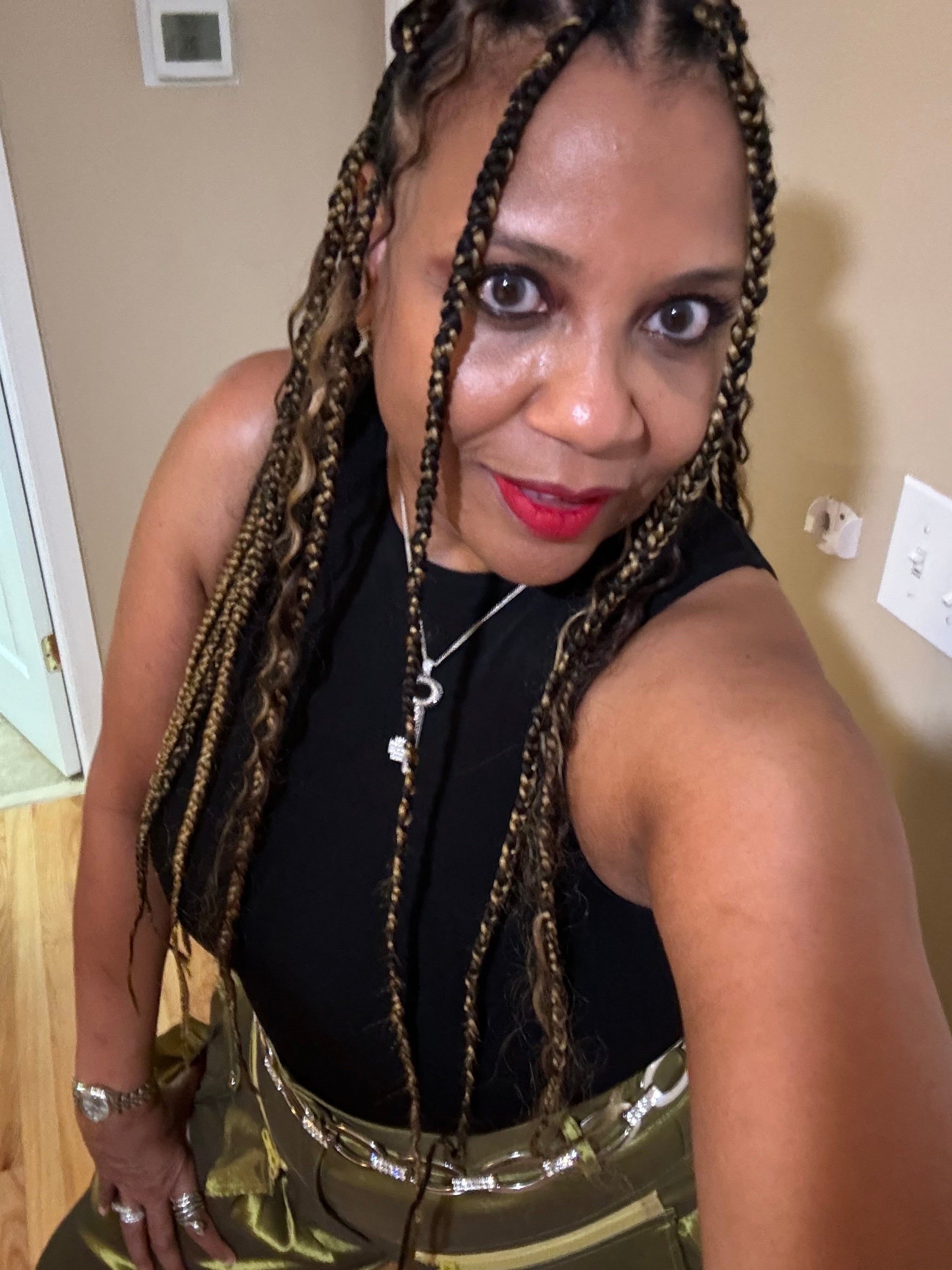 Eva, Herbs and Oils CEO wears blond highlighted braids with bright red lipstick a black tank and green shiny pants.  She is sitting and looking into the camera.