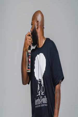 Man with brown skin, bald head, and large beard holding a bottle of The Tonic and spraying it on his beard while wearing an Herbs and Oils Tee Shirt