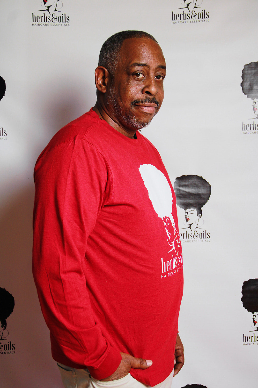 Picture of David Burke standing in front of an Herbs and Oils backdrop wearing a red herbs and oils tee shirt with a fresh haircut after using herbs and oils products on his hair and beard.