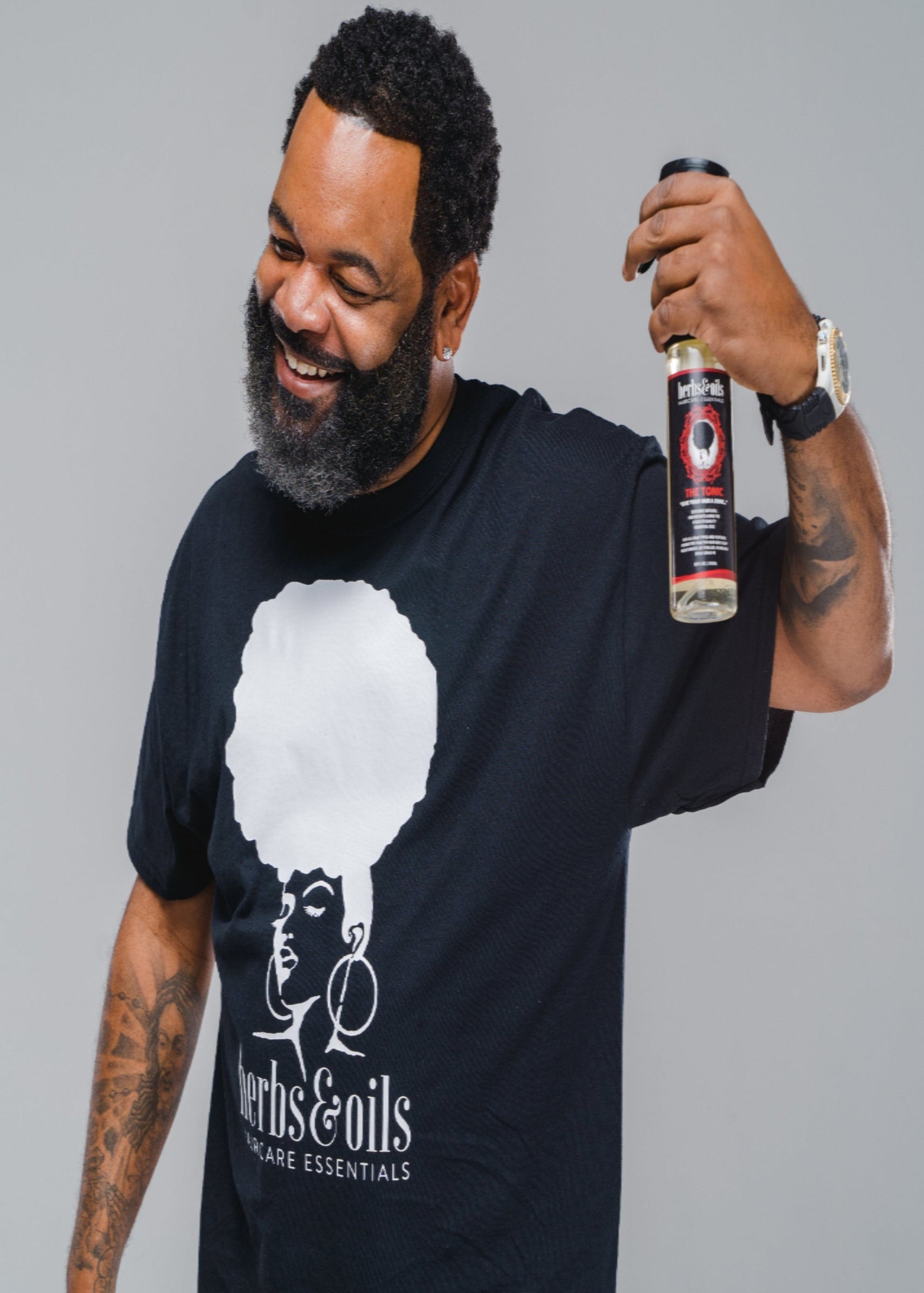 Black man in an Herbs and Oils tee shirt holding a bottle of Herbs and Oils, The Tonic, with a salt and pepper beard and clean haircut and full head of hair, showing how he uses The Tonic on his hair and beard to retain his hair as he ages.
