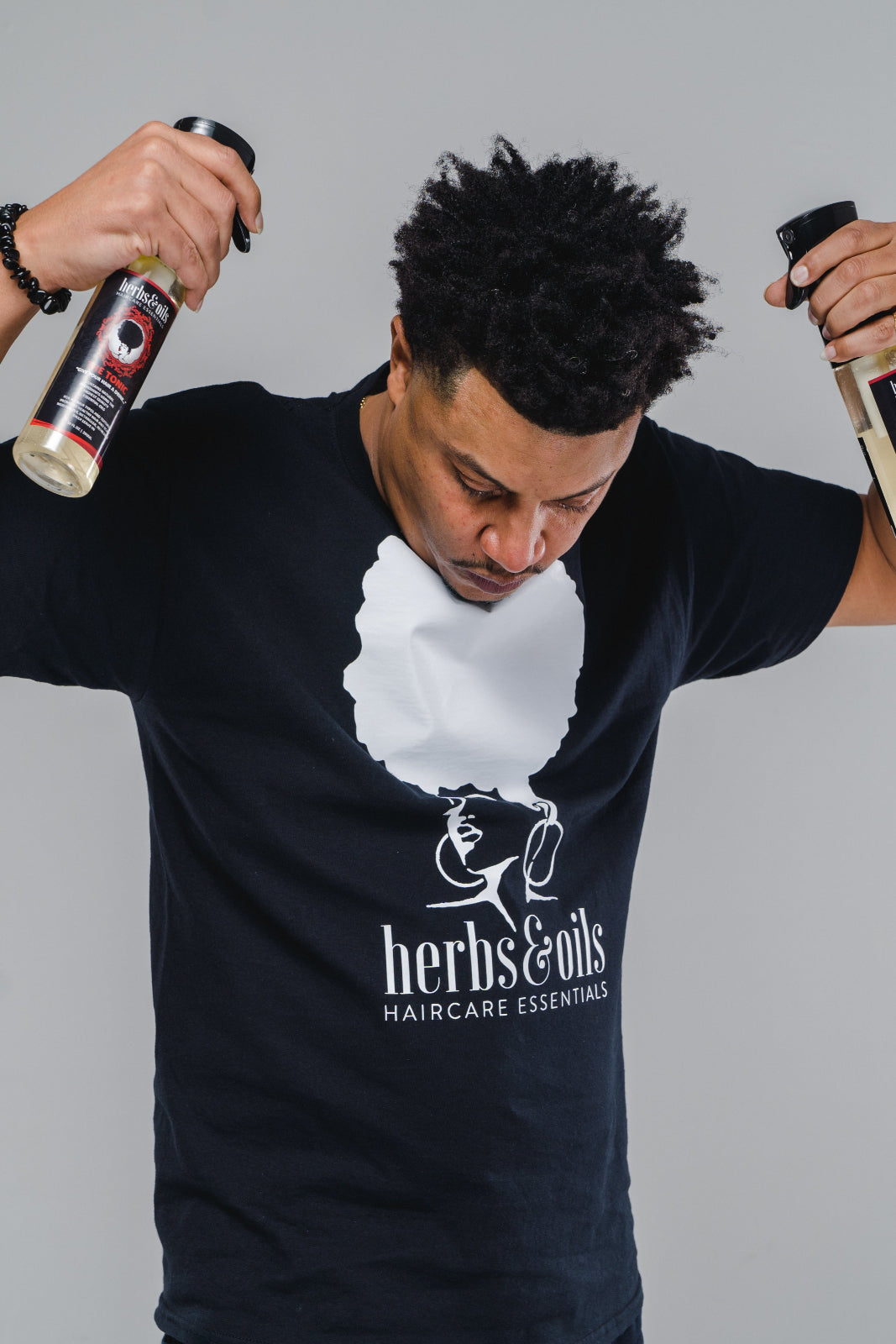 Black man with textured hair, wearing an Herbs and Oils tee shirt and holding two bottles of Herbs and Oils Haircare, The Tonic as if he is spraying them both