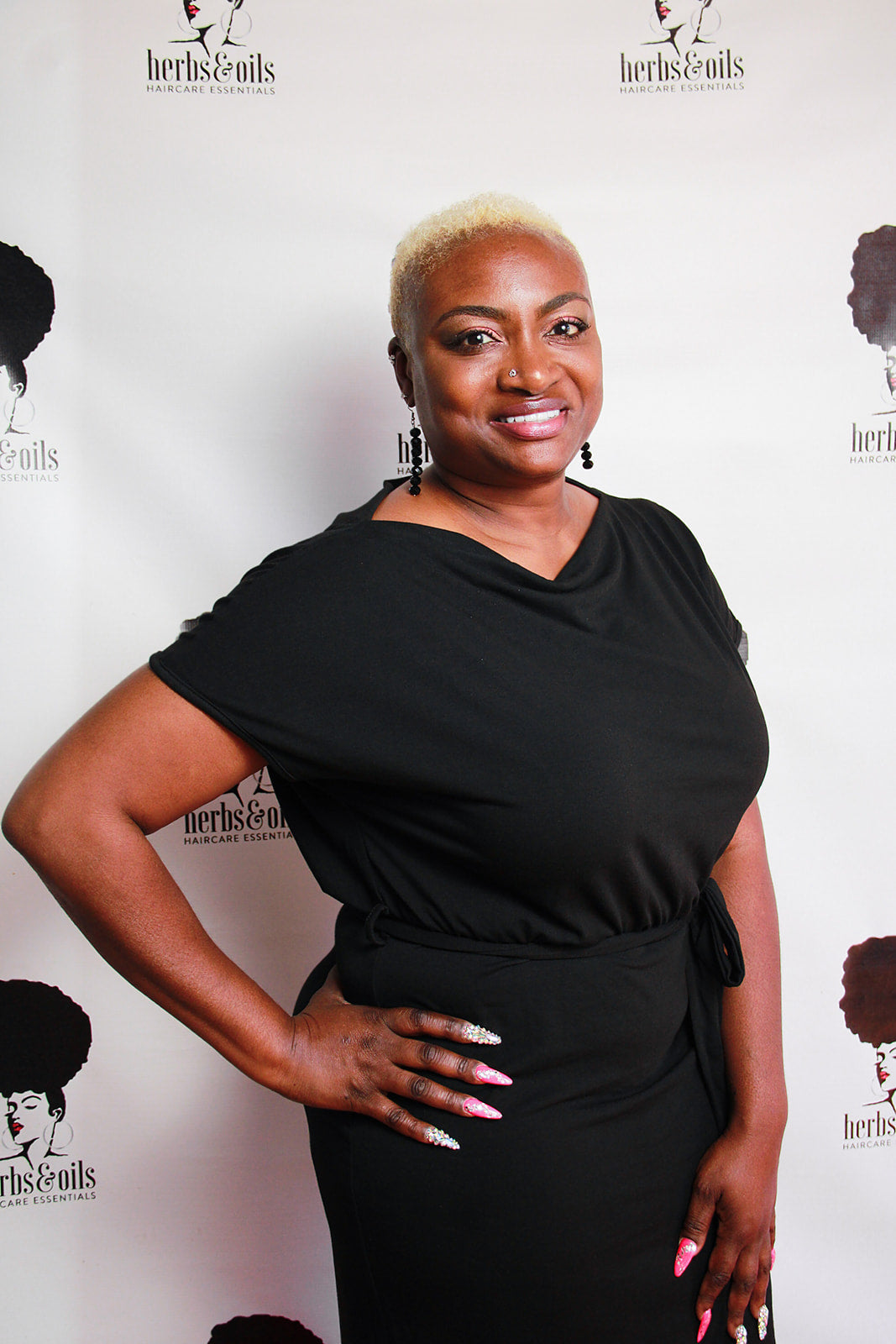 Mature lady with a short cut dyed blond in a natural style.  Used The Tonic, Curly Cocktail, and Last Call Finishing Oil to style color treated hair.  Standing in front of an herbs and oils banner showing her beautiful nails