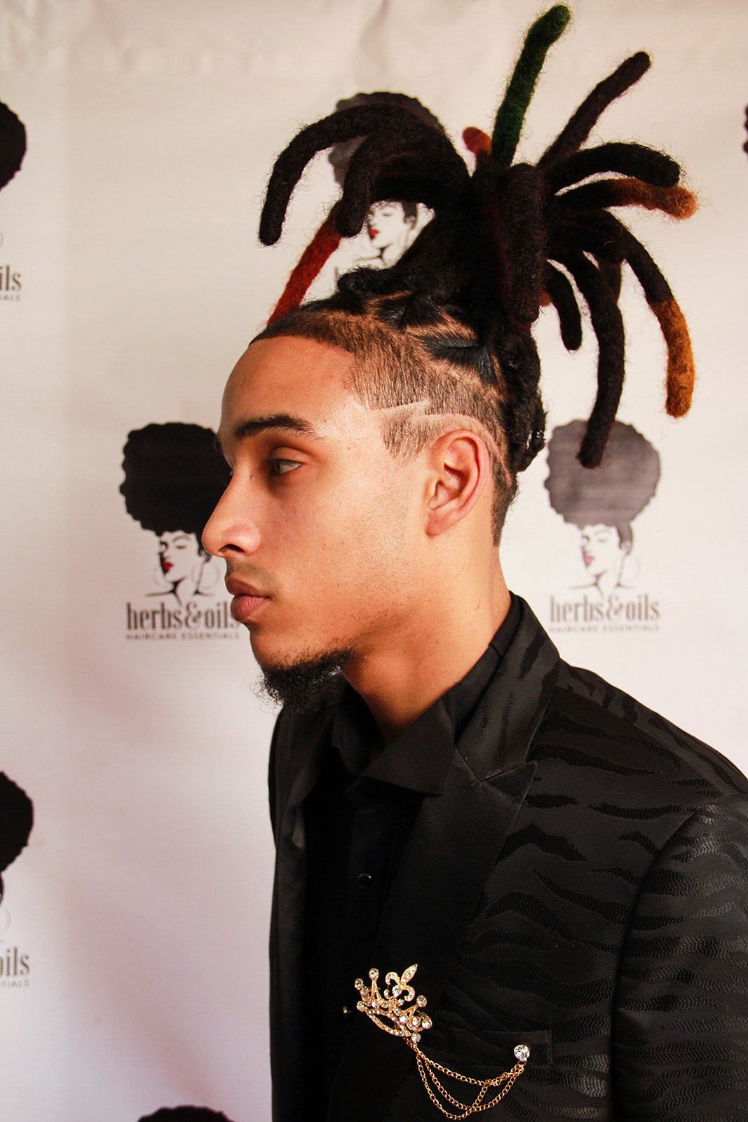 Young man with fresh loc retwist with wicks that are color treated at the ends.  Side profile in front of an herbs and oils banner